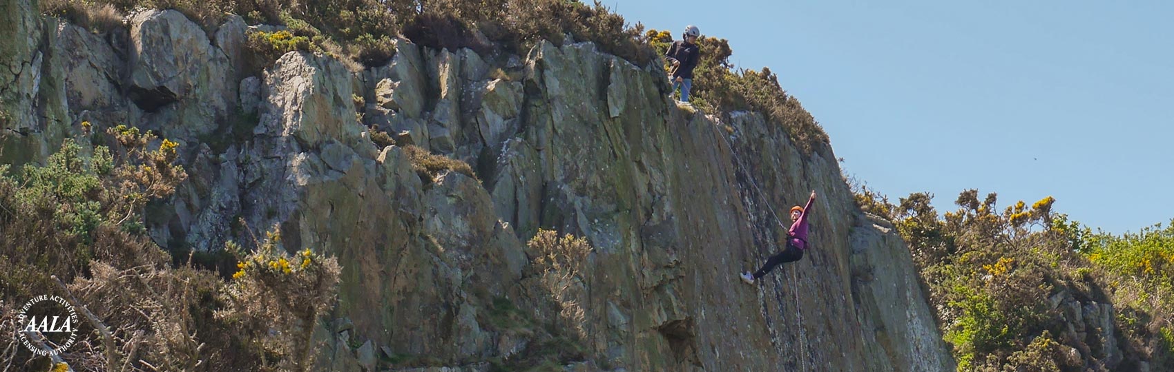 Woman Abseiling down a large cliff face, waves at the camera