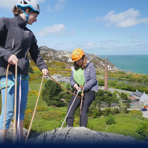 A girl is abseiling down a large cliff face for the first tim as a guilde supports her above
